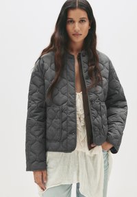 Grå kviltad jacka med rund halsringning, framsida med knappar och en ficka. Bärs över en ljuskrämfärgad topp som kompletterar denimjeansen.