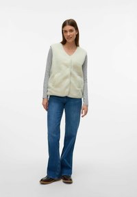 Gilet évasé vert clair en laine avec un col en V, fermeture par boutons et deux poches latérales, porté sur une chemise à manches longues grise et un jean évasé bleu.