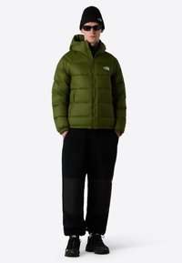 Grön puffjacka med dragkedja fram och huva, svarta byxor med texturerad yta, svarta sneakers och tillbehör som en mössa och solglasögon.