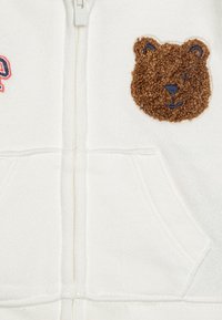 Baltā kapucīna jaka ar brūnu pūkainu lāča pārslu un izšūtām detaļām. Mīksta auduma, ar priekšējo kabatu un izšūtām akcentiem.