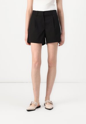 Femme portant un short noir sur mesure, un haut blanc et des ballerines beiges pointues à double bride, debout sur un fond blanc.