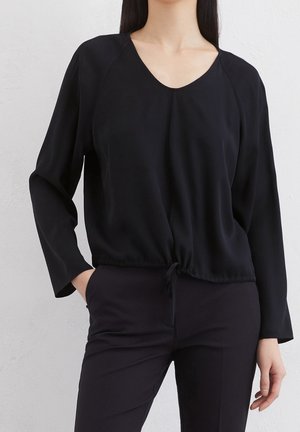Kobieta ubrana w czarną bluzkę z długim rękawem z dekoltem w serek i sznurkiem w talii, w połączeniu z czarnymi dopasowanymi spodniami na tle jednolitej ściany.