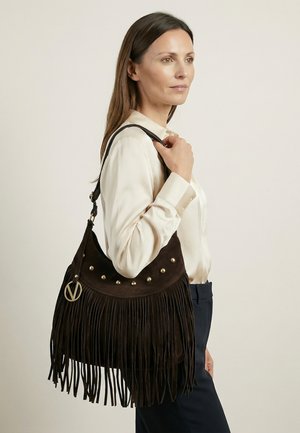 Femme portant un chemisier crème et un pantalon sombre, tenant un grand sac à franges marron foncé avec des clous dorés posé sur son épaule.