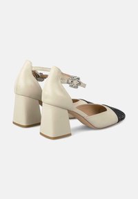 Scarpe con tacco a blocco in pelle beige con una punta intrecciata nera e una cinghia decorativa alla caviglia con un accent gemma trasparente.