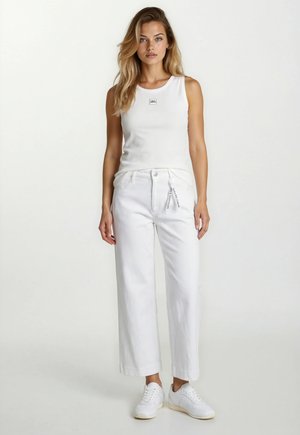 Young woman with long hair wearing a white sleeveless top, high-waisted wide-leg white pants, and white sneakers, standing against a plain background.