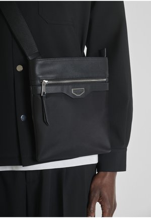 Borsa a tracolla nera con parte superiore testurizzata e cerniera argentata indossata sopra un abito nero sul lato destro della persona.