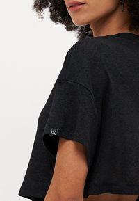 T-shirt nera a maniche corte e cropped, realizzata in tessuto morbido. Presenta un piccolo tag con il logo sulla manica, evidenziando un design minimalista.