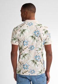 Polo a maniche corte di colore beige con una stampa floreale tropicale in blu e verde. Tessuto morbido, colletto a costine e vestibilità rilassata.