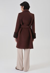 Cappotto di lana marrone con cintura, polsini rifiniti in pelliccia, design sartoriale e taglio a mezza lunghezza, abbinato a pantaloni beige a gamba larga.