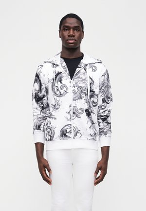 Młody mężczyzna stoi, ubrany w białą bluzę z kapturem z czarno-szarymi abstrakcyjnymi wzorami oraz białe spodnie na jednolitym tle.