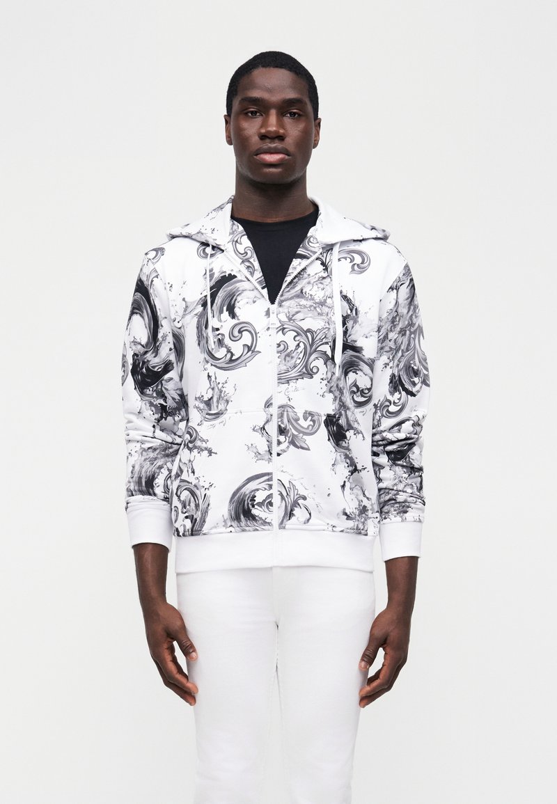 Νεαρός άνδρας στέκεται φορώντας λευκό φούτερ με μαύρους και γκρι αφηρημένους στροβίλους και λευκό παντελόνι μπροστά από απλό φόντο.