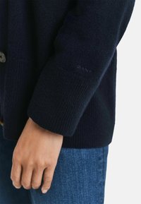 Marineblaue Strickjacke mit gerippten Bündchen, ausgestattet mit einem Knopfverschluss und dezentem Branding am Ärmel, kombiniert mit Jeans.