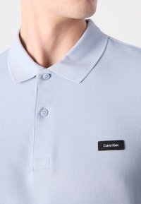 Polo shirt bleu clair en tissu texturé, avec un col classique, deux boutons et un patch logo Calvin Klein noir.