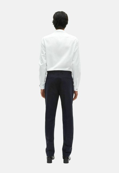 Mężczyzna stojący plecami, ubrany w białą koszulę z długim rękawem, ciemnoniebieskie spodnie i czarne eleganckie buty na jednolitym tle.