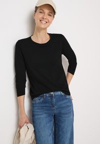 Schwarzes Langarm-T-Shirt mit rundem Halsausschnitt, kombiniert mit hellblauen Jeans. Das Model hält eine beige Jacke und trägt eine passende Mütze.
