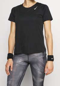 Camiseta atlética negra de manga corta y cuello redondo; presenta un pequeño logo plateado de Nike en el hombro izquierdo. Combinada con leggings estampados.