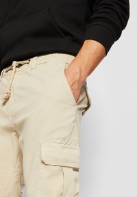 Person trägt beige Cargohose, Hand in der Tasche, schwarzes Langarmshirt vor einfarbigem Hintergrund.