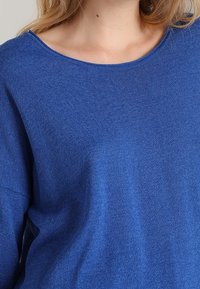 Maglione blu a maglia con un ampio scollo rotondo, morbido al tatto e con spalle scese. Il tessuto appare leggermente testurizzato, offrendo una vestibilità rilassata.