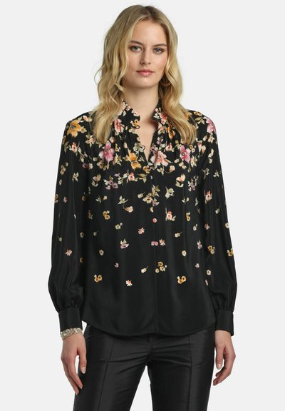 Femme blonde portant un chemisier noir à manches longues avec des motifs floraux roses, jaunes et blancs, associée à un pantalon noir, debout devant un fond blanc.