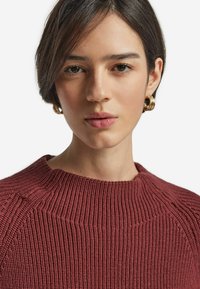 Rostfärgad stickad tröja med ribbad struktur och hög halsringning. Guldörhängen i form av ringar kompletterar looken. Enfärgad utan mönster.