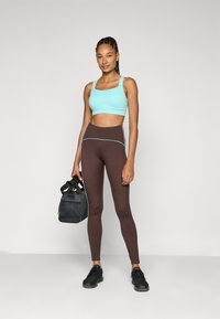 En türkis sports-bh med brede stropper, sammen med høyt liv brune leggings med lyseblå piping, og en svart treningsveske.