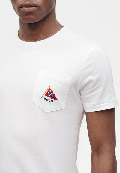 Κοντινό πλάνο ενός ατόμου που φοράει λευκό T-shirt με μικρό τσεπάκι στο στήθος με κεντημένο λογότυπο Polo σε κόκκινο, μπλε και κίτρινο.