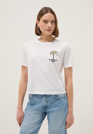 Mujer con camiseta blanca recortada con una palmera y texto "Palm Beach Florida", combinada con jeans azul claro sobre un fondo neutro.