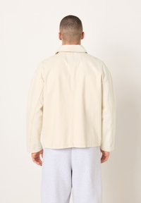 Übergroße Jacke in Cremefarbe mit Kragen, sichtbaren Nähten und geradem Schnitt. Getragen über hellgrauen Jogginghosen.