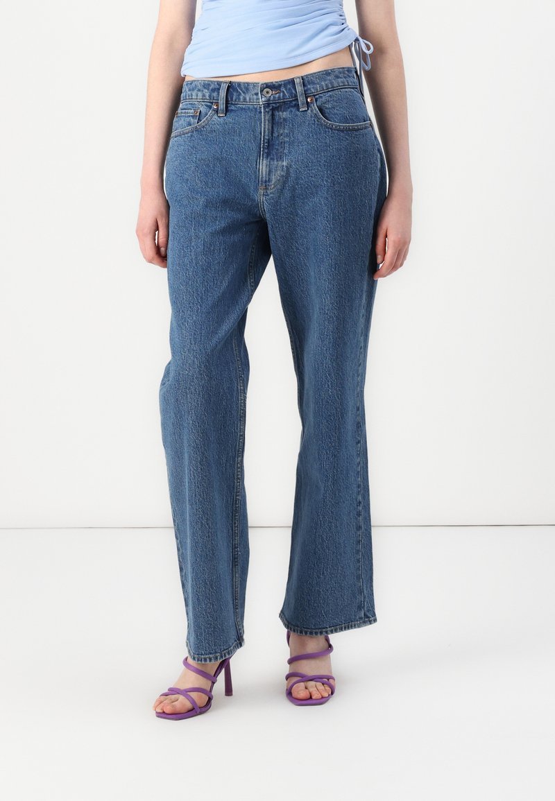 High-waisted blue denim jeans with a relaxed fit, featuring five pockets, belt loops, and a light texture. Paired with purple strappy sandals.