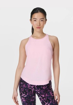 Jonge vrouw met donker haar in een staart, draagt een lichtroze mouwloos topje en paarse legging met bloemmotief tegen een effen lichte achtergrond.