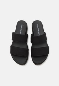 Black slide sandals with two wide fabric straps and a smooth rubber sole. The brand name "Calvin Klein Jeans" is printed on the insole.