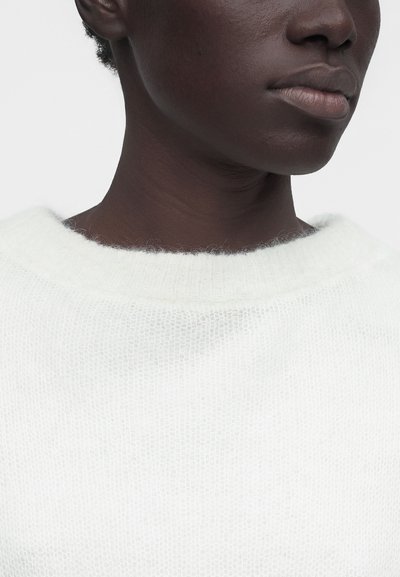 Άτομο με σκουρόχρωμο δέρμα που φοράει ένα μαλακό, χνουδωτό λευκό πουλόβερ, με εμφανή λαιμό, γραμμή σιαγόνας και μέρος των χειλιών πάνω σε απλό φόντο.