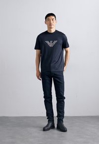 Camiseta de algodón negra con un logotipo gráfico en el pecho, combinada con jeans oscuros y botas de tobillo negras, de pie frente a un fondo liso.