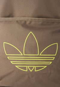 Ruda tekstūra pasižyminti audinio medžiaga, kurioje yra didelis, ryškus geltonas „Adidas“ logotipas su trimis lapais ir trimis horizontaliais dryžiais.
