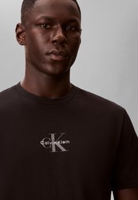 Mladý muž s tmavou pletí, který má na sobě černé tričko Calvin Klein s logem uprostřed na hrudi, na jednobarevném šedém pozadí.