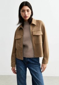 Giacca in suede marrone con due tasche anteriori, ampio colletto e chiusura con zip, indossata sopra un maglione beige a coste e jeans blu scuro.