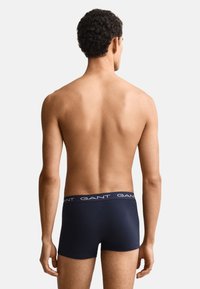Mann mit kurzen lockigen Haaren, von hinten gezeigt, trägt dunkelblaue GANT Boxershorts vor einem einfarbigen hellen Hintergrund.
