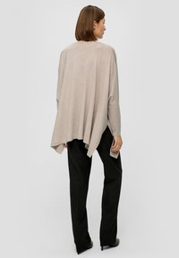 Beige, oversized gebreide top met een gedrapeerde achterkant en lange mouwen, gecombineerd met zwarte rechte broek en hoge hakken. Glad textuur.