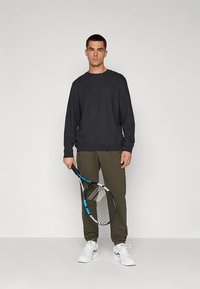 Sudadera gris oscuro con cuello redondo, pantalones ajustados color verde oliva y zapatillas deportivas blancas; sosteniendo una raqueta de tenis con detalles azules.