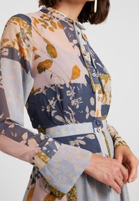 Robe à manches longues avec col, présentant un design en patchwork dans des teintes de bleu, rose et vert, avec des motifs floraux et des détails de boutons.