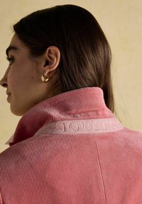 Tume juustega naine, kes kannab kuldseid poolkuu kõrvarõngaid ja roosa sametjaki, mille tagakaelusel on tikandiga sõna "JOULES".