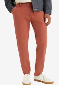 Chinos couleur rouille en coton, avec une coupe slim, des poches latérales et une texture lisse. Associés à des baskets blanches.