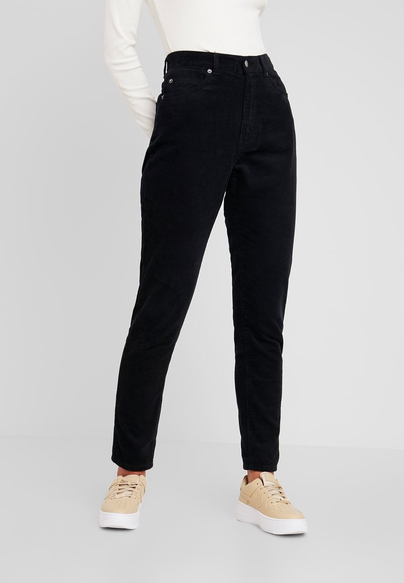 Personne portant un pantalon en velours noir, des baskets beiges et un haut blanc à manches longues, debout devant un fond gris clair uni.