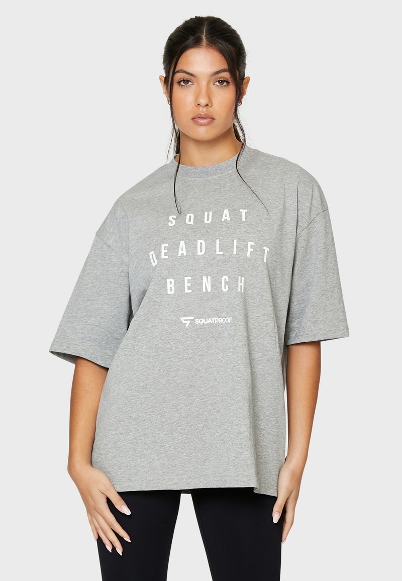 Harmaa oversize-t-paita, joka on valmistettu puuvillasta. Paidan etuosassa on rohkea valkoinen grafiikka, jossa lukee "SQUAT DEADLIFT BENCH" kauluksen alla.