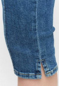 Pantalons capri en denim de couleur bleu délavé, avec une fente sur le bas et des détails de couture contrastants jaunes.