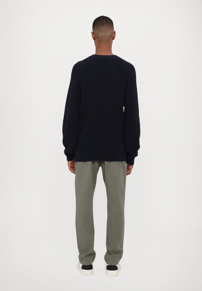 Pull en maille marine avec un motif texturé et un col rond, associé à un pantalon vert clair. Les chaussures comportent un détail noir.