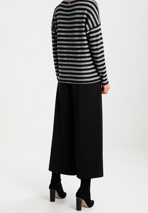 Persona vistiendo un suéter de rayas grises y negras, pantalones negros de pierna ancha y botines negros de tacón alto, vista desde atrás.