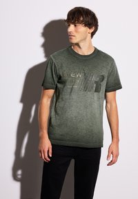 Tmavě zelené bavlněné tričko s texturovaným povrchem. Obsahuje grafický potisk v světlejším odstínu. Kulatý výstřih a krátké rukávy.