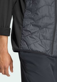 Veste noire avec un panneau matelassé gris foncé, dotée d'une fermeture éclair et de manches en texture douce. Les matériaux semblent légers et durables.