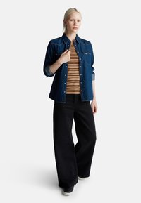 Jeansowa kurtka z jasnoniebieskimi wewnętrznymi mankietami, pasek w brązowo-kremowe paski, czarne spodnie szerokie u dołu i czarne sneakersy, prezentowane na białym tle.